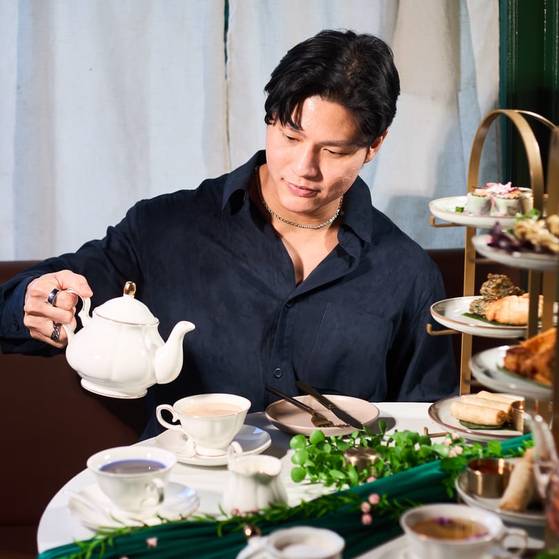 A man pours tea from a white teapot into a cup at a table with Thai appetizers.