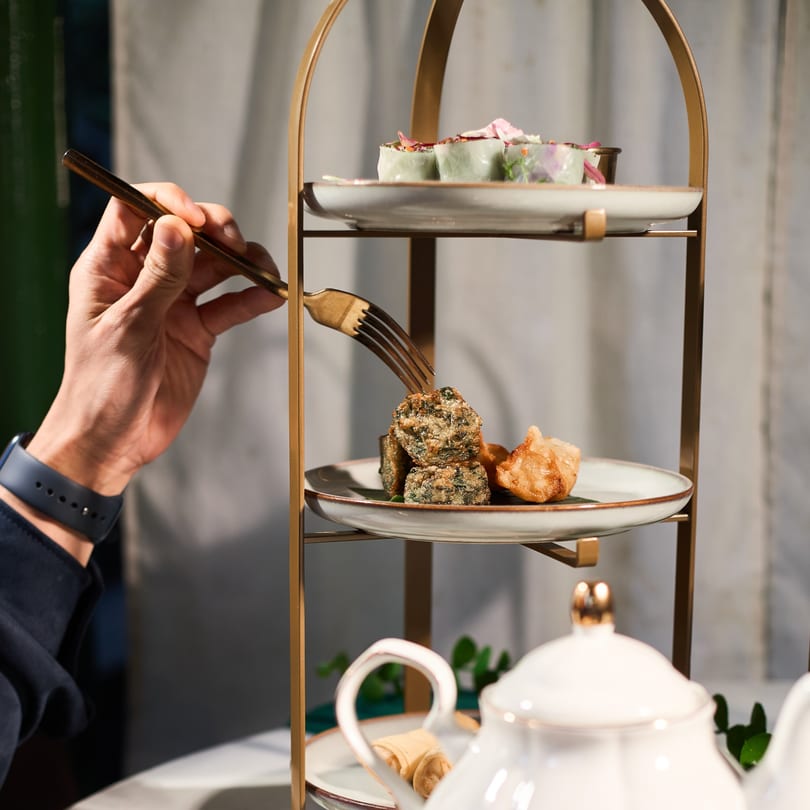 A hand uses a fork to pick up a piece of fried Thai food from a tiered serving tray.