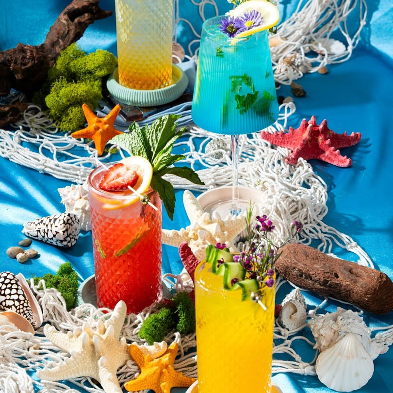Four colorful Thai cocktails with garnishes, surrounded by seashells, starfish, and fishing net on blue.