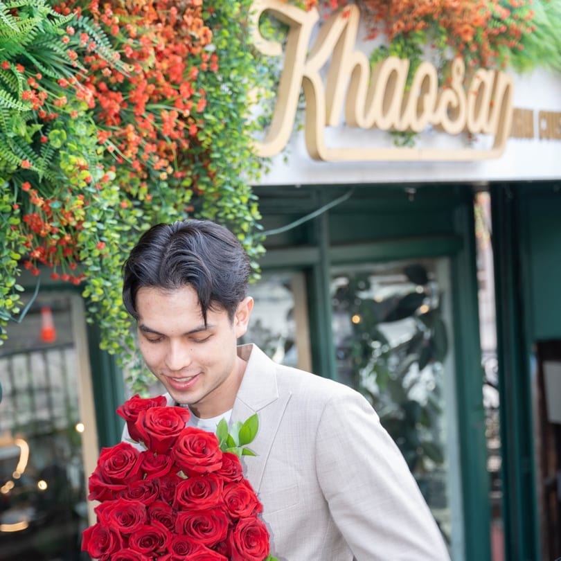 A man in a light suit holds a large bouquet of red roses outside Khaosan Thai restaurant.