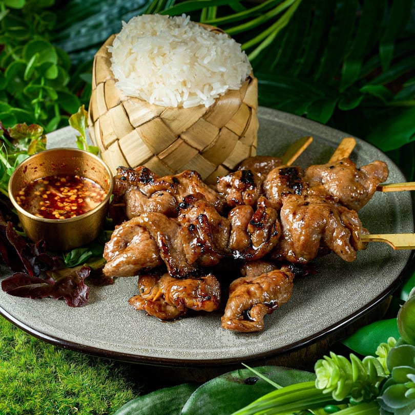 Grilled pork skewers with sticky rice in a bamboo basket and chili dipping sauce on a plate.