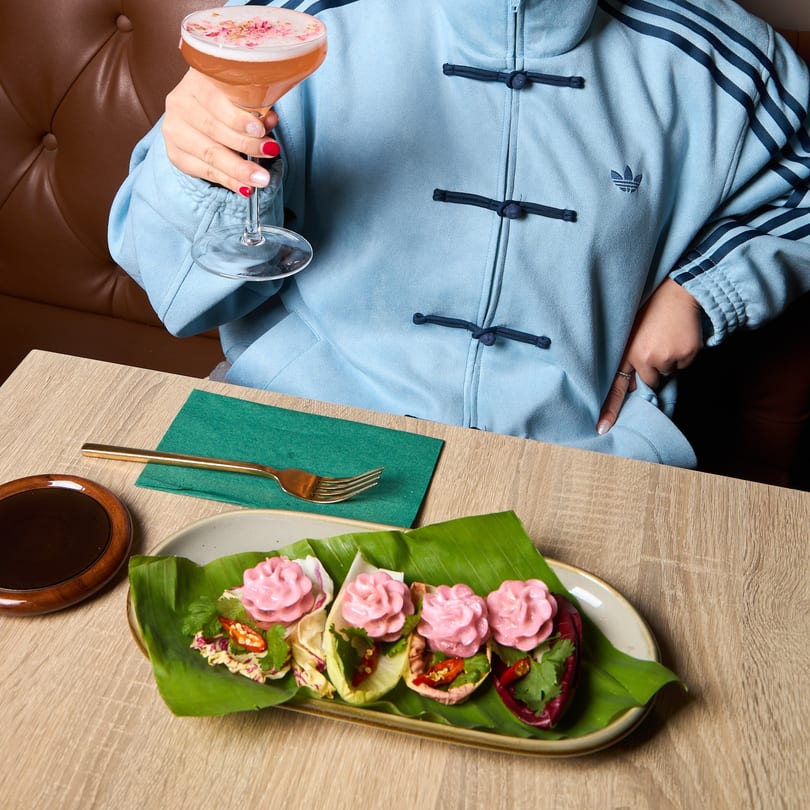 Person in blue jacket holding a pink cocktail, with Thai appetizers on banana leaves on a table.