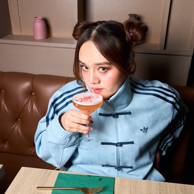 A woman in a blue Adidas jacket sips a pink cocktail with foam and petals at Khaosan.
