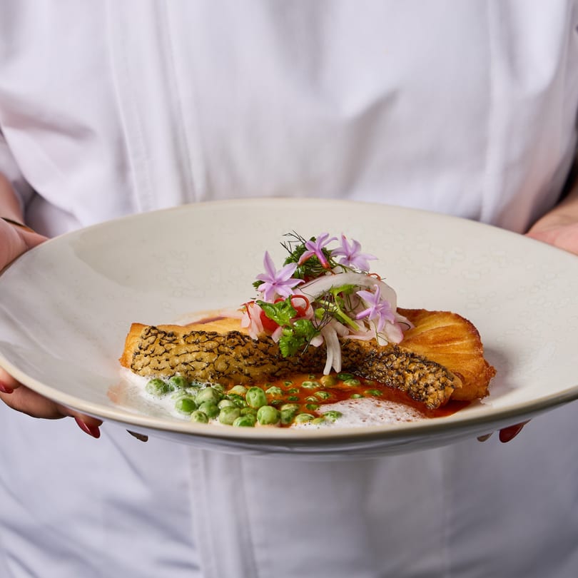 Chef holding a plate of crispy-skinned fish with peas, red sauce, and purple flowers.