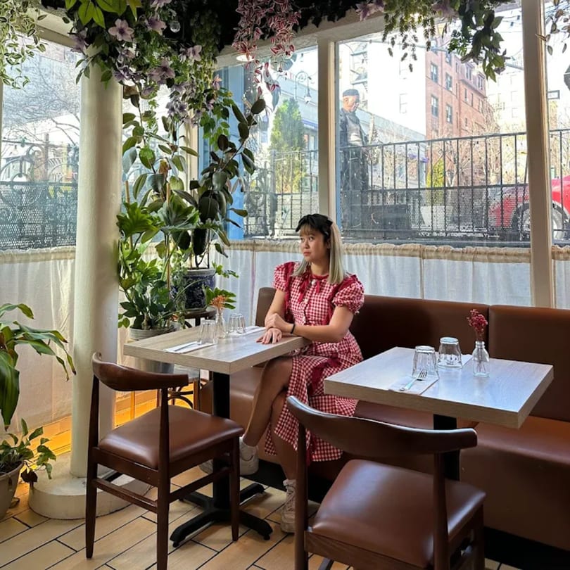 A woman in a red gingham dress sits at a table in a bright, plant-filled Khaosan restaurant.