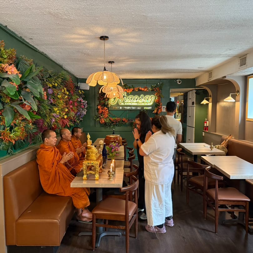 Monks in orange robes and people praying in a Thai restaurant with green walls and floral decor.