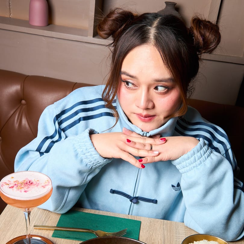 A woman with space buns and a blue jacket sits at a table with a pink cocktail.