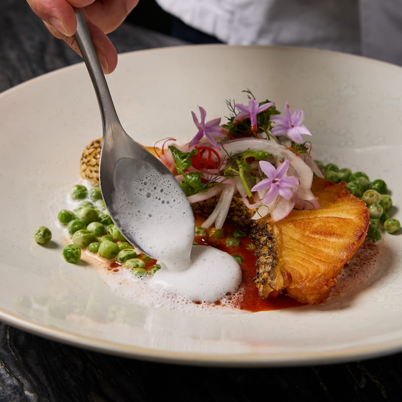A chef spoons foam onto a Thai dish of crispy fish, peas, and edible flowers.